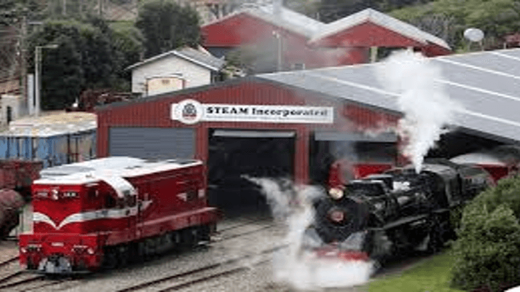 Red diesel locomotive and black steam locomotive emerging from a red storage building