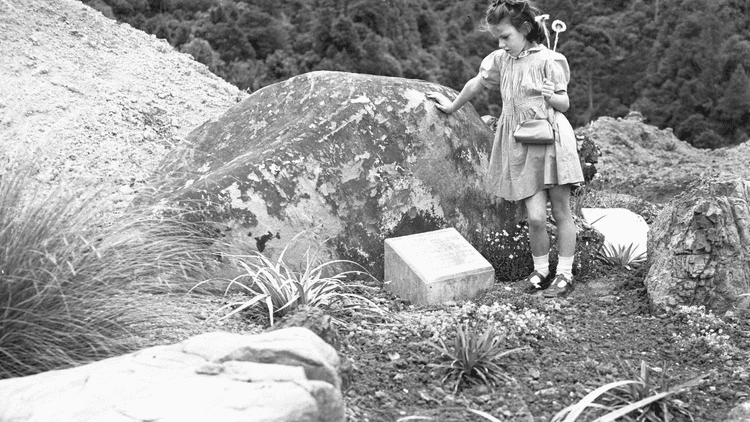 Black and white photo of a young girl with her hand resting against a rock holding gardening tools starting down at the ground