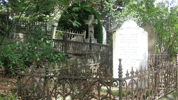 Photo of a tombstone surrounded by an aged fence with another tombstone in the background, surrounded by trees. 
