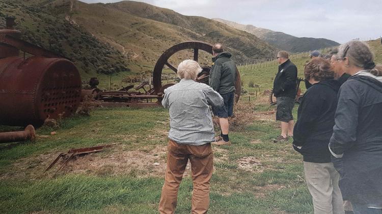 Photo of a group of people listening to a talk with hills in the background with some of the machinery laying on the grass. 